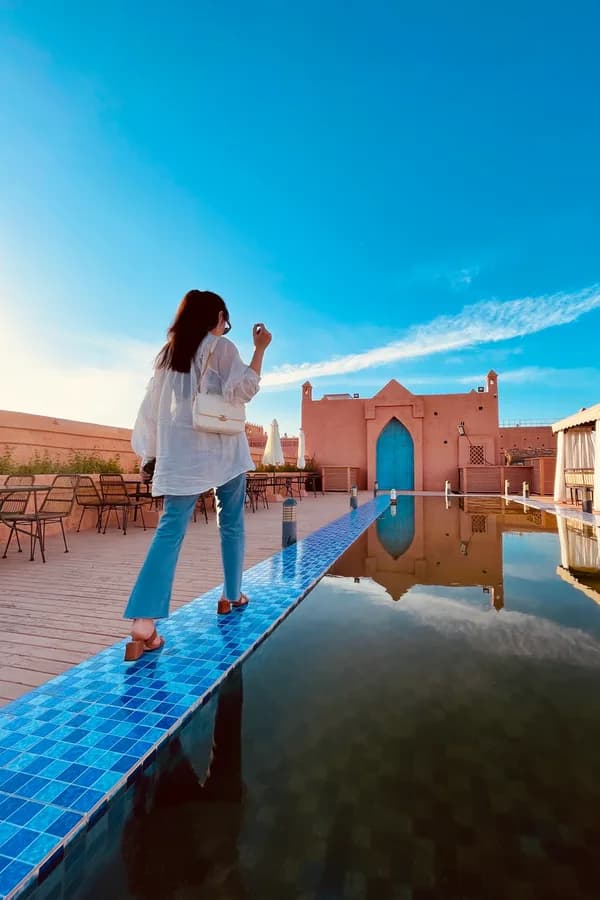 A traveller walking along the blue-tiled edge of a rooftop pool in Morocco, in front of a terracotta wall with an arched blue door