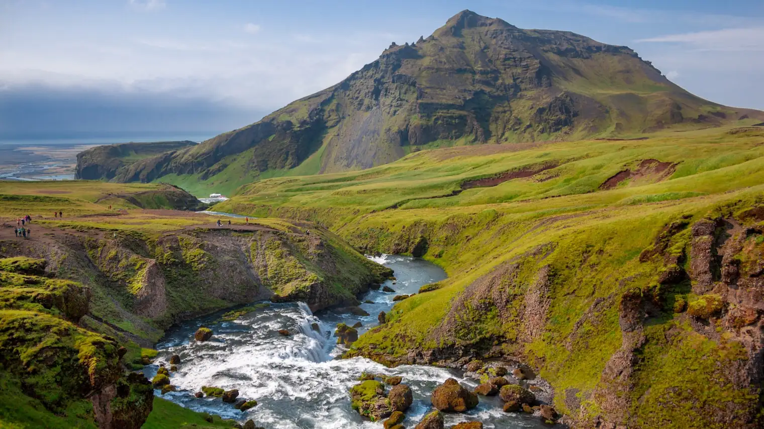 A river cutting through bright green Icelandic highland, with walkers on the ridge path above the falls