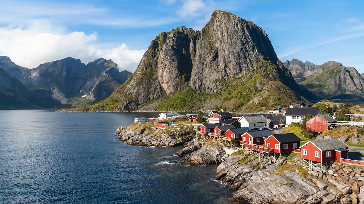 Red wooden fishing cabins on stilts below a steep granite peak on a Norwegian fjord in summer