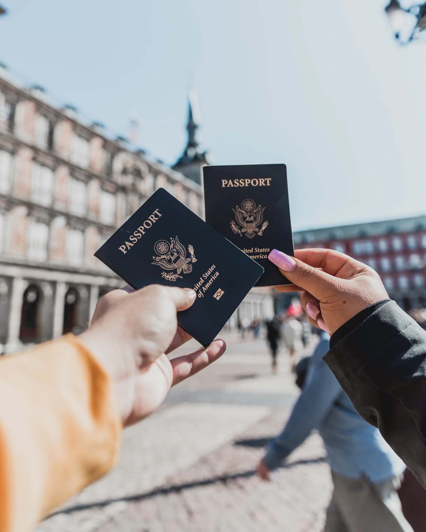 Dos manos sosteniendo pasaportes estadounidenses en alto en una plaza europea, un viajero haciendo una pausa en su viaje