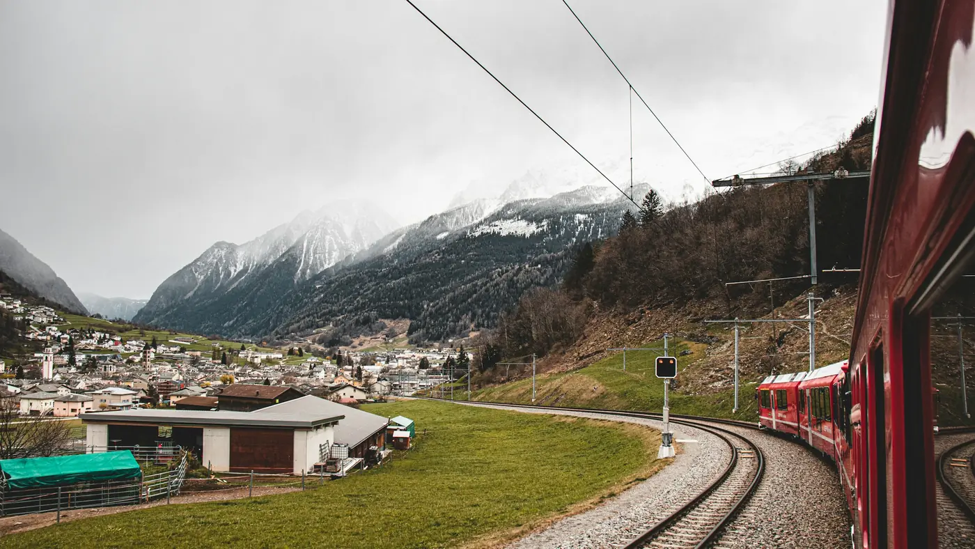 A red train curving past an Alpine village near St Moritz, Switzerland, with snow-dusted peaks behind