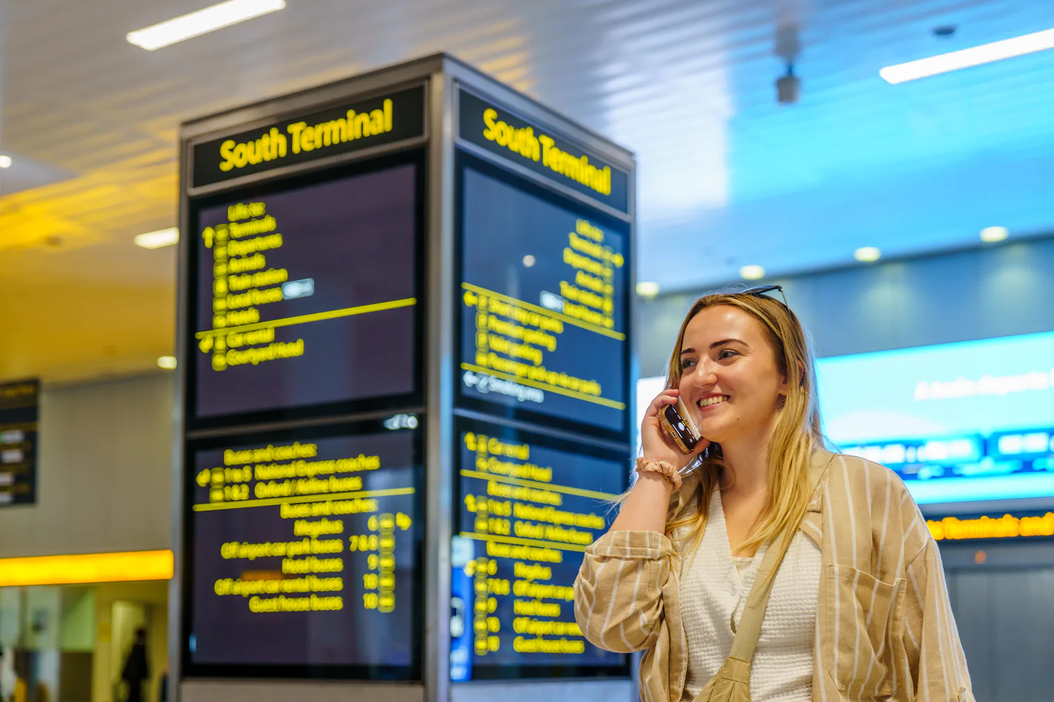 Viajera sonriendo al teléfono delante del panel informativo de una terminal de aeropuerto