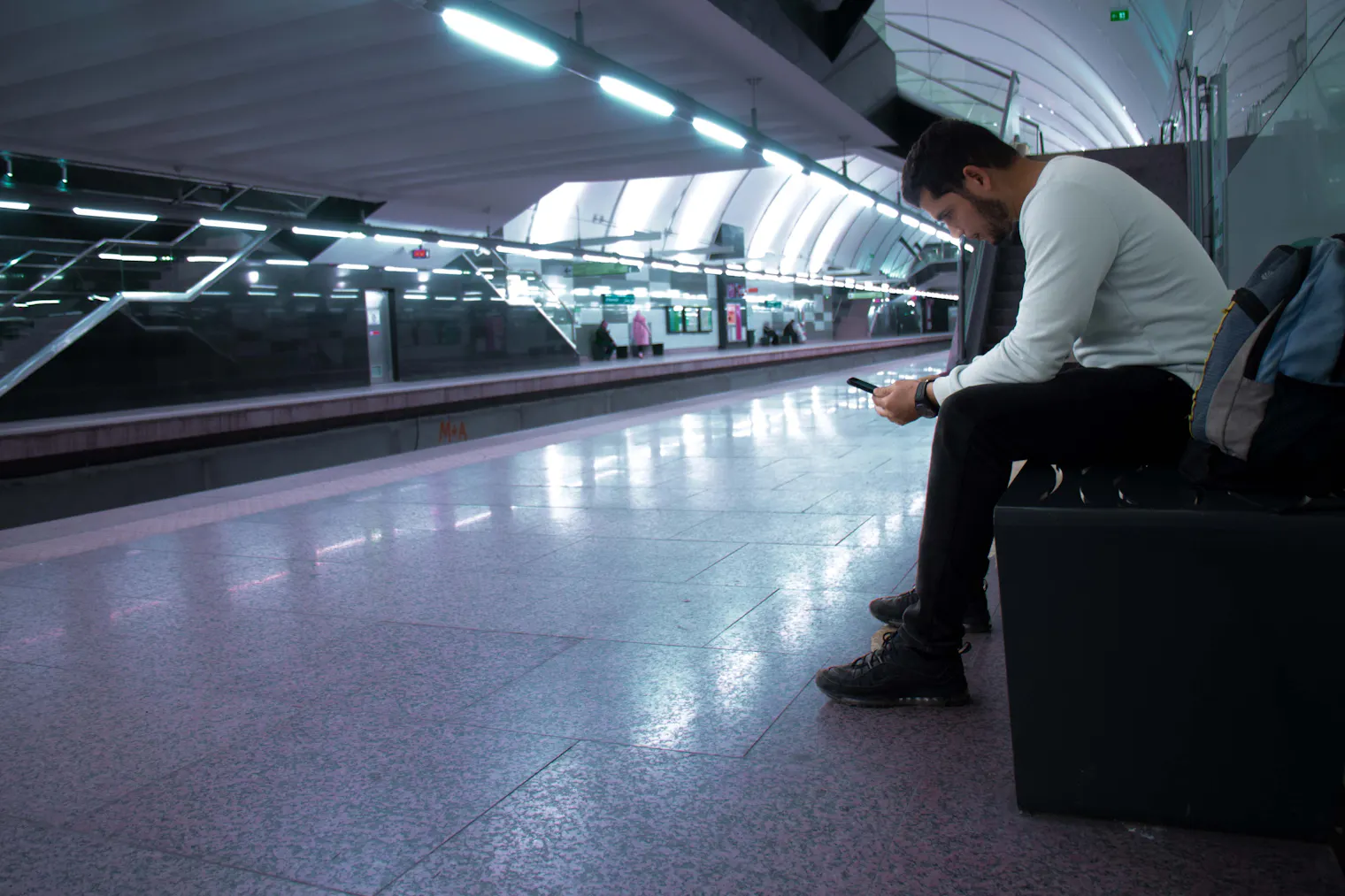 Traveller with a backpack sitting alone on an empty metro platform, waiting for something to load on their phone