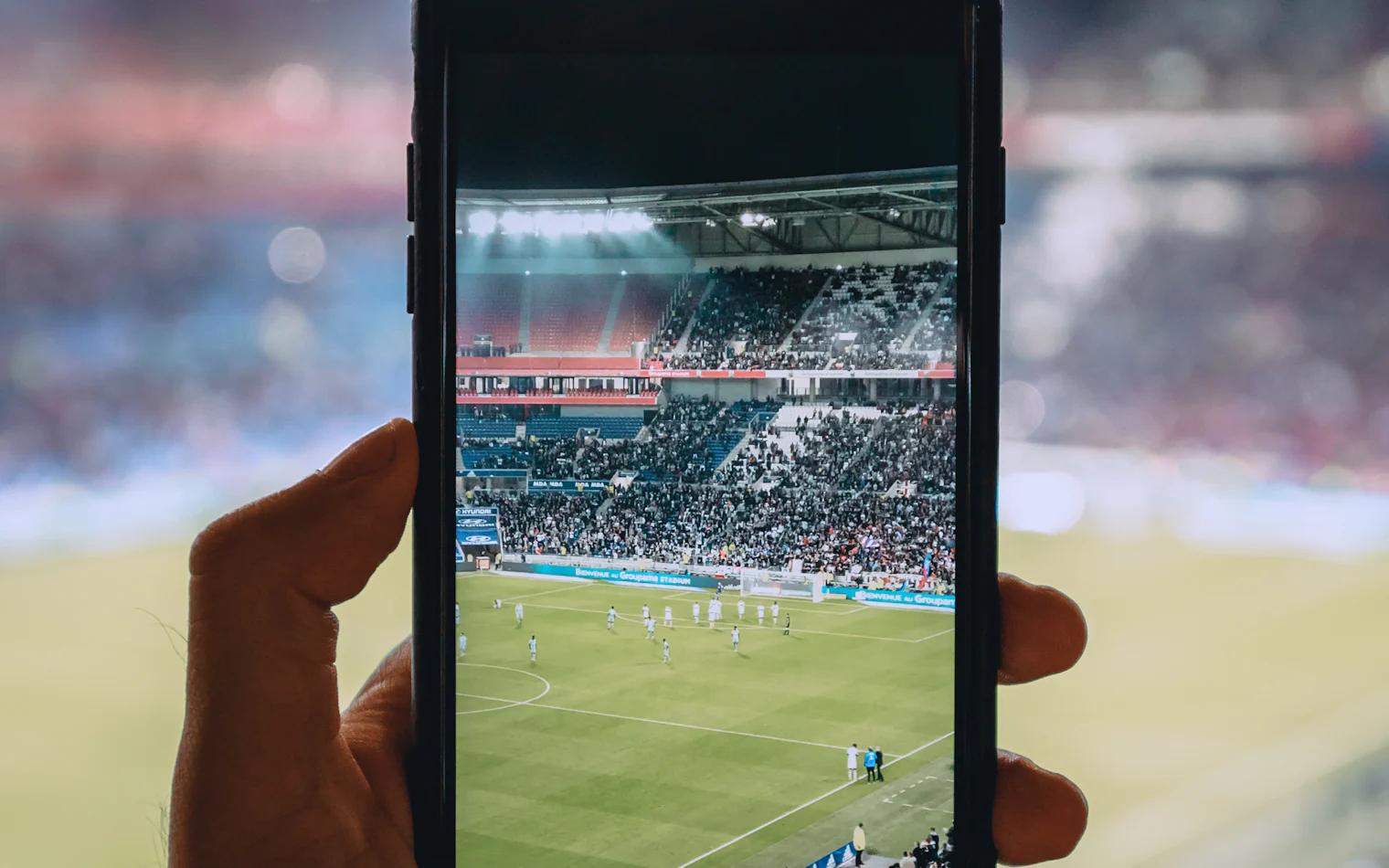 Fan holding up a smartphone showing a live football match inside a packed stadium